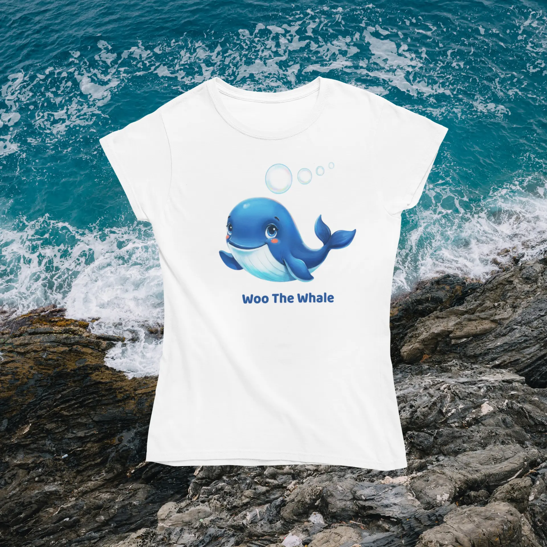 White T-shirt with a blue cartoon whale graphic set against a rocky shoreline and crashing waves; shirt text reads 'Woo The Whale' at the bottom.